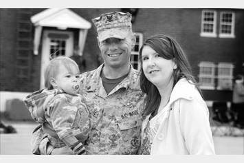 dad in his army uniform holding his baby and posing with his wife. they are standing in front of a house
