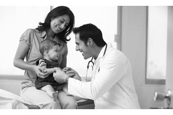 Mother holding baby on her lap in a doctor's office, while a physician examines the baby.