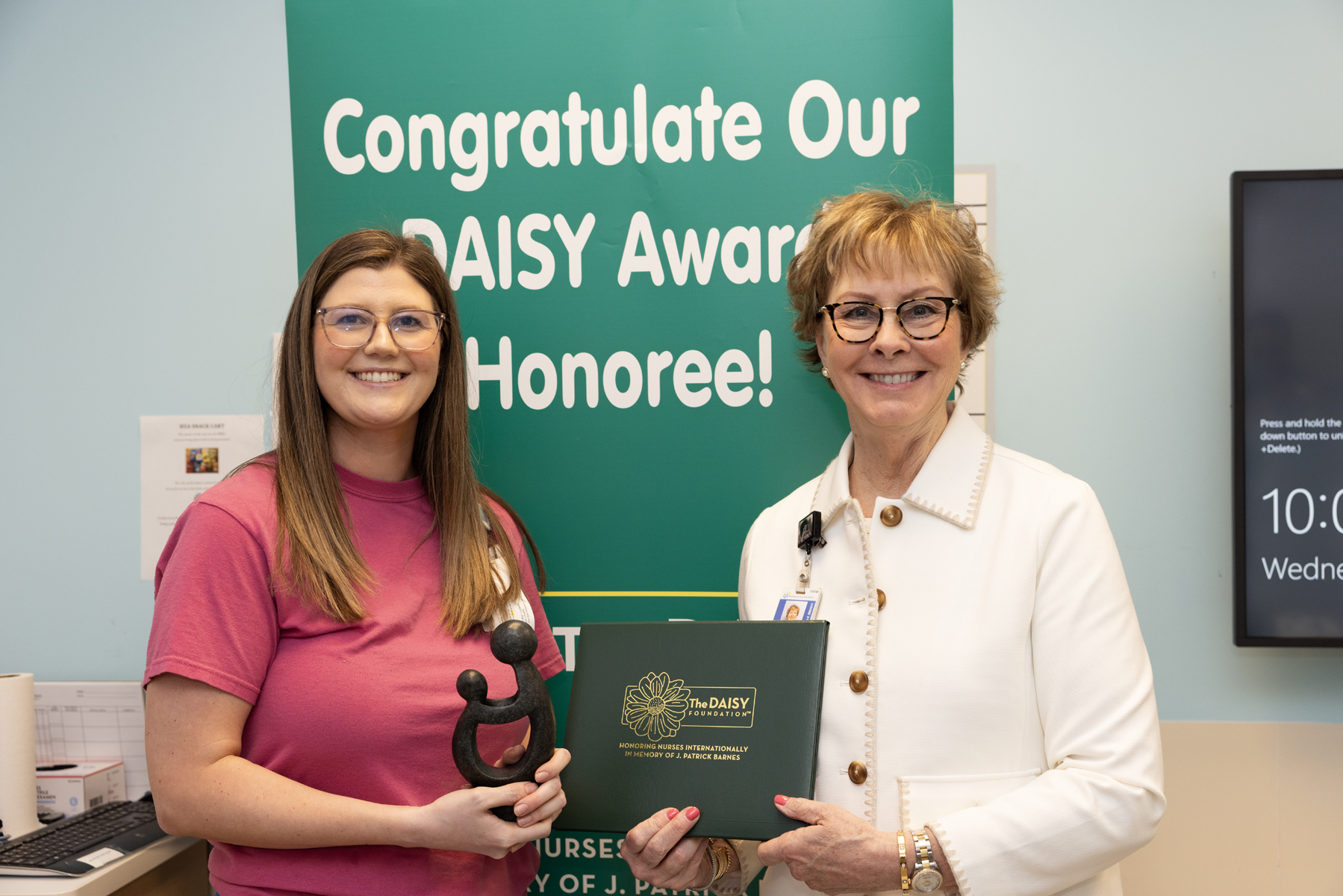 A nurse smiles while receiving the DAISY Award, holding the signature statue. She stands next to a hospital leader who presents the award certificate. Behind them is a congratulatory banner recognizing the honoree for exceptional nursing care.